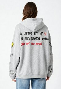 Pelēka oversized kapuce ar auklu. Satur melnu un sarkanu tekstu, saules grafiku un nolietota sirds dizainu uz piedurknes.