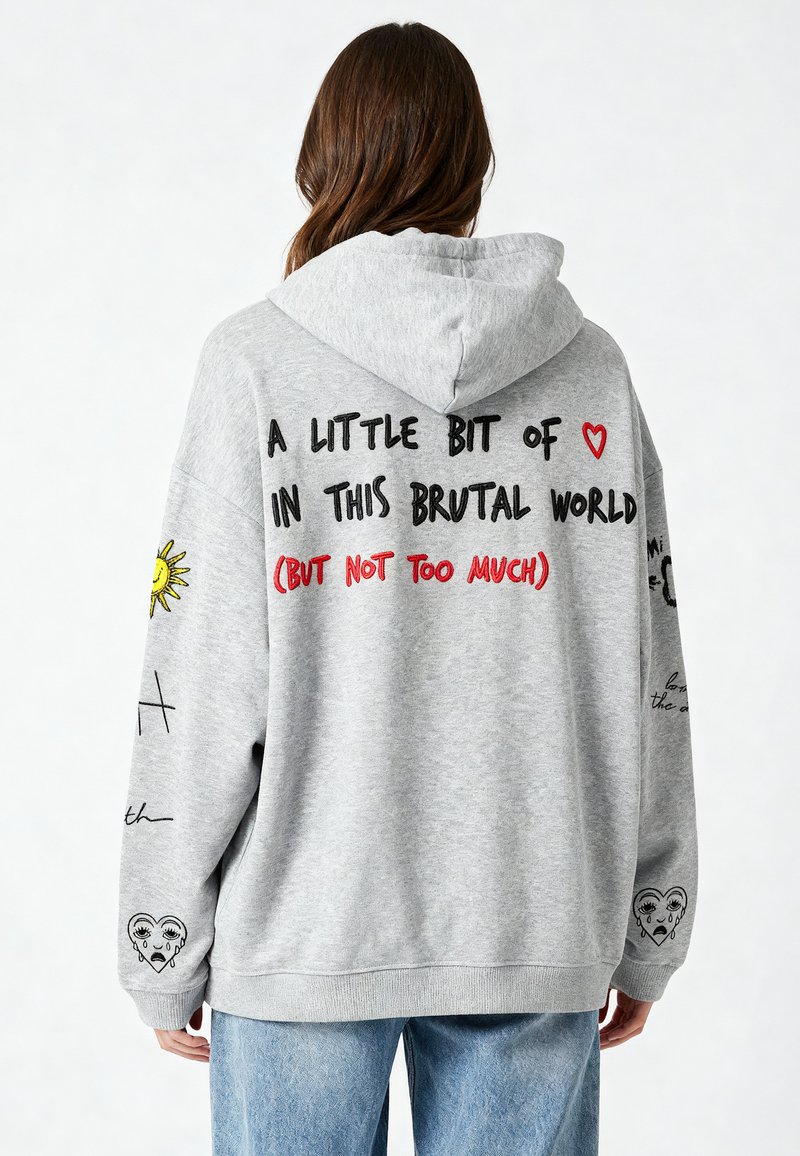 Pelēka oversized kapuce ar auklu. Satur melnu un sarkanu tekstu, saules grafiku un nolietota sirds dizainu uz piedurknes.