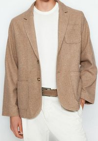 Uomo che indossa un blazer di lana beige sopra una camicia bianca e pantaloni bianchi con una cintura beige, con una mano in tasca, in piedi contro uno sfondo neutro.