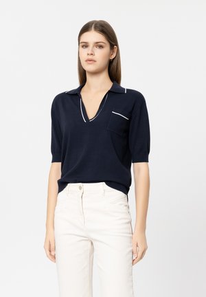 Frau mit glattem braunem Haar trägt ein marineblaues Kurzarmoberteil mit weißem Rand und hellfarbene, hochtaillierte Hosen vor einfachem Hintergrund.