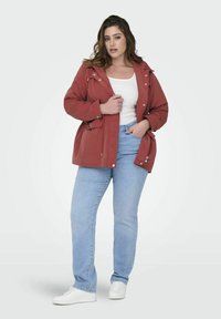 Giacca utilitaria rossa con cappuccio, chiusura a bottoni e tasche laterali, indossata sopra una maglietta bianca e jeans azzurri chiari, abbinata a sneaker bianche.