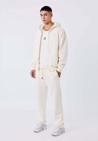 Joven de pie vestido con una sudadera color crema, pantalones de chándal a juego, camiseta gráfica blanca y zapatillas blancas sobre un fondo liso.