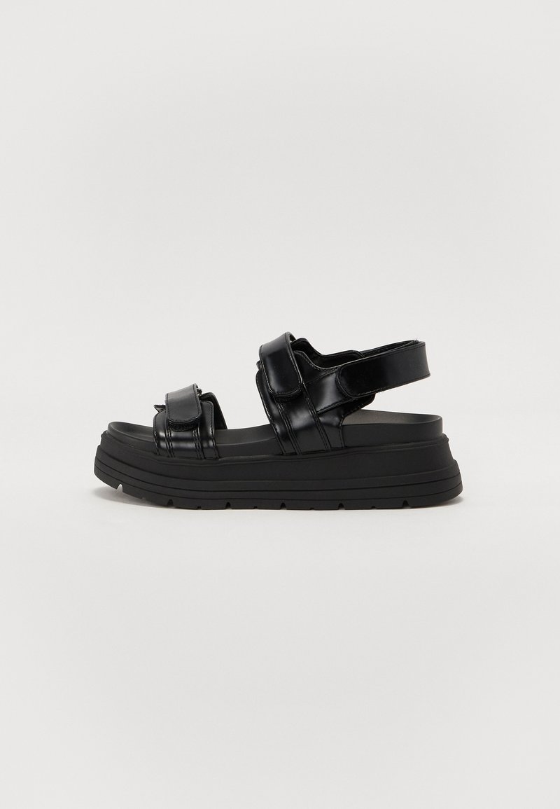 Sandalias de plataforma negras hechas de material brillante, con correas anchas y una suela acolchada con dibujo para agarre. Correa trasera ajustable.