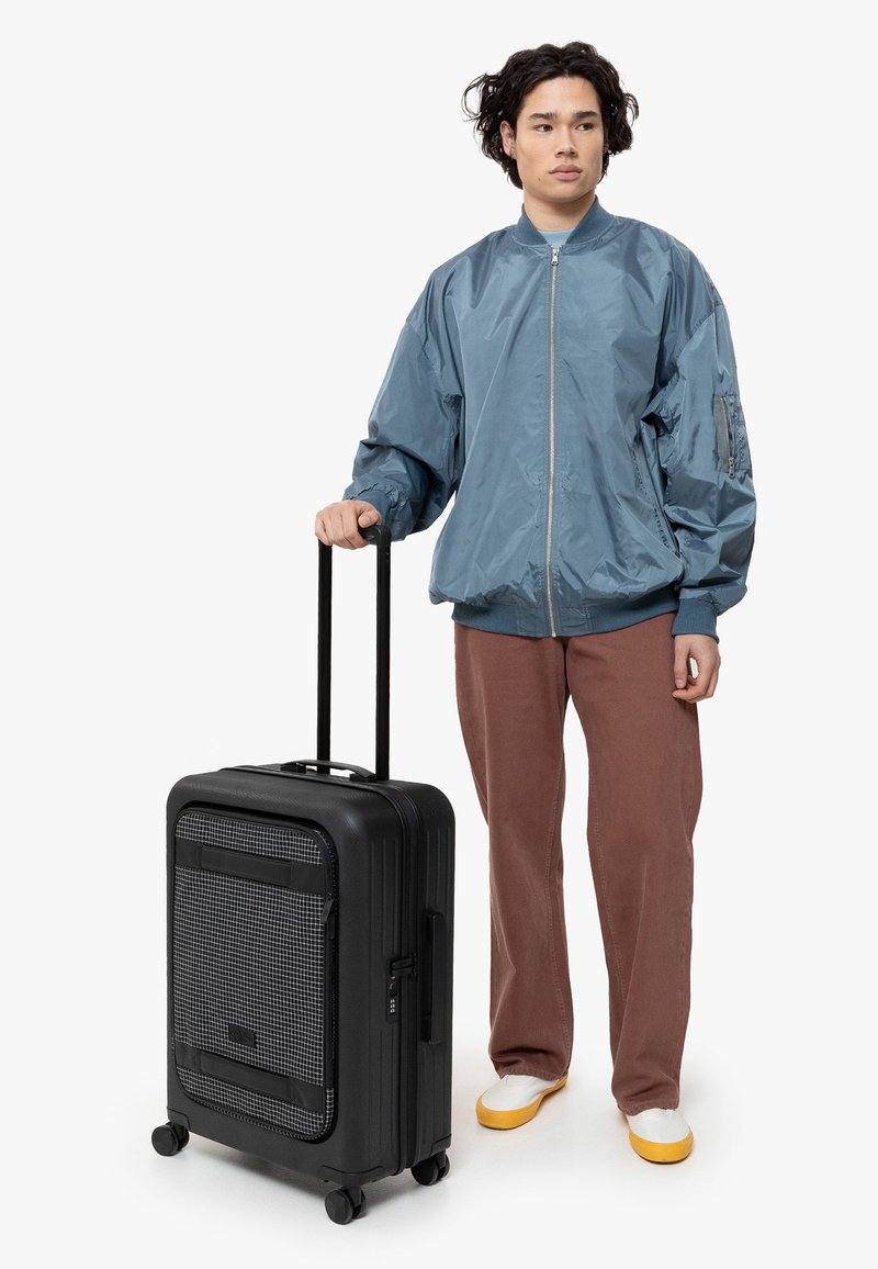 Hartschalenkoffer in Schwarz mit einem Gittermuster auf der Vorderseite, ausziehbarem Griff und vier Rädern. Modell trägt eine blaue Bomberjacke und braune Hose.