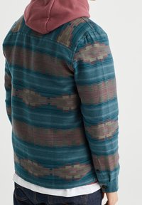 Mönstrad skjorta med en teal-färgad bas och geometriska mönster i rosa och grönt, långa ärmar och en avslappnad passform, lager-på-lager över en hooded tröja.