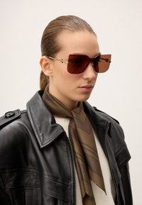 Brune oversized solbriller med guld accenter, båret af en person i en sort læderjakke og et mønstret brunt tørklæde.