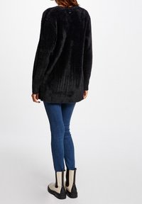 Žena oblečená v huňatém černém svetru s dlouhým rukávem, tmavě modrých skinny džínách a béžovo-černých kotníkových botách, stojící s otočením zády.