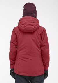 Veste isolée rouge avec capuche, design de dos sans couture et poignets texturés. Portée avec un bonnet sombre et des gants noirs.