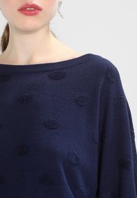 Marinblå stickad tröja med off-shoulder halsringning, dekorativt mönster av texturerade bollar genom hela plagget och med ett mjukt, lätt tyg.