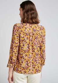 Blouse florale moutarde avec des motifs violets, roses et verts. Manches longues avec des poignets retroussés et un détail plissé à l'arrière. Texture de tissu lisse.