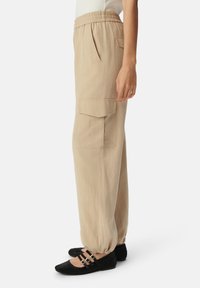 Beige cargobroek met een elastische tailleband, zijzakken en een relaxte fit. Zwarte flats met drie gespsmuigingen.