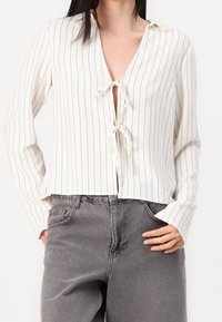 Femme portant un chemisier crème à rayures verticales à manches longues avec deux liens à l'avant et un jean gris délavé taille haute, les mains dans les poches.