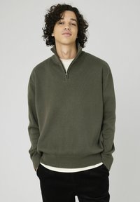 Un pull zippé vert olive en tissu texturé avec des poignets côtelés, porté sur une chemise couleur crème, associé à un pantalon en velours côtelé noir.