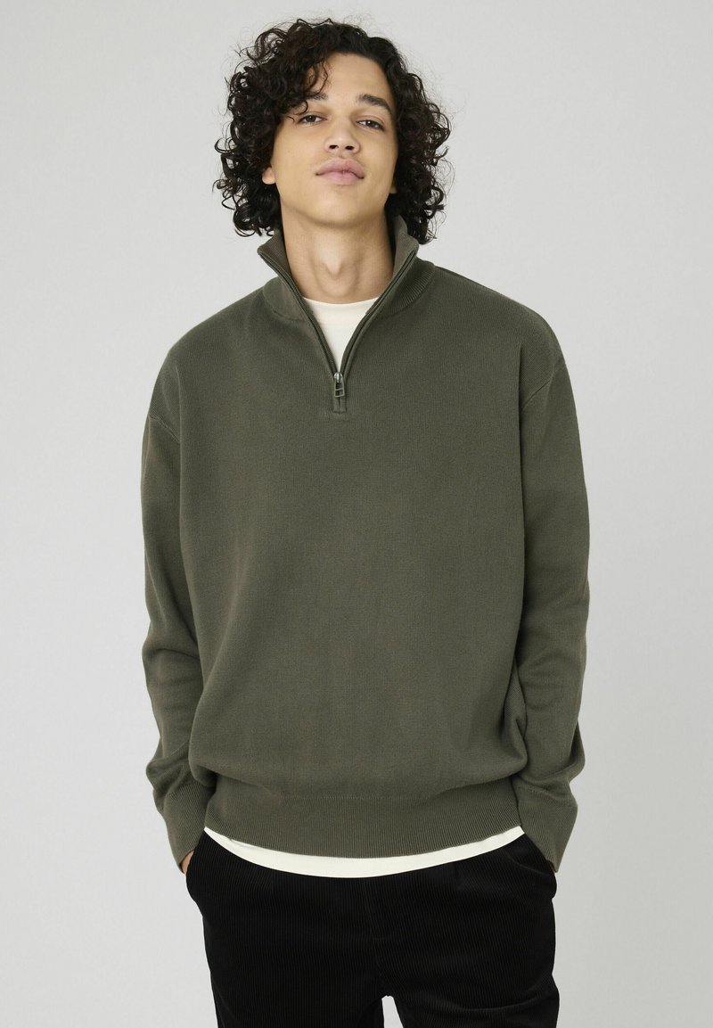 Un pull zippé vert olive en tissu texturé avec des poignets côtelés, porté sur une chemise couleur crème, associé à un pantalon en velours côtelé noir.