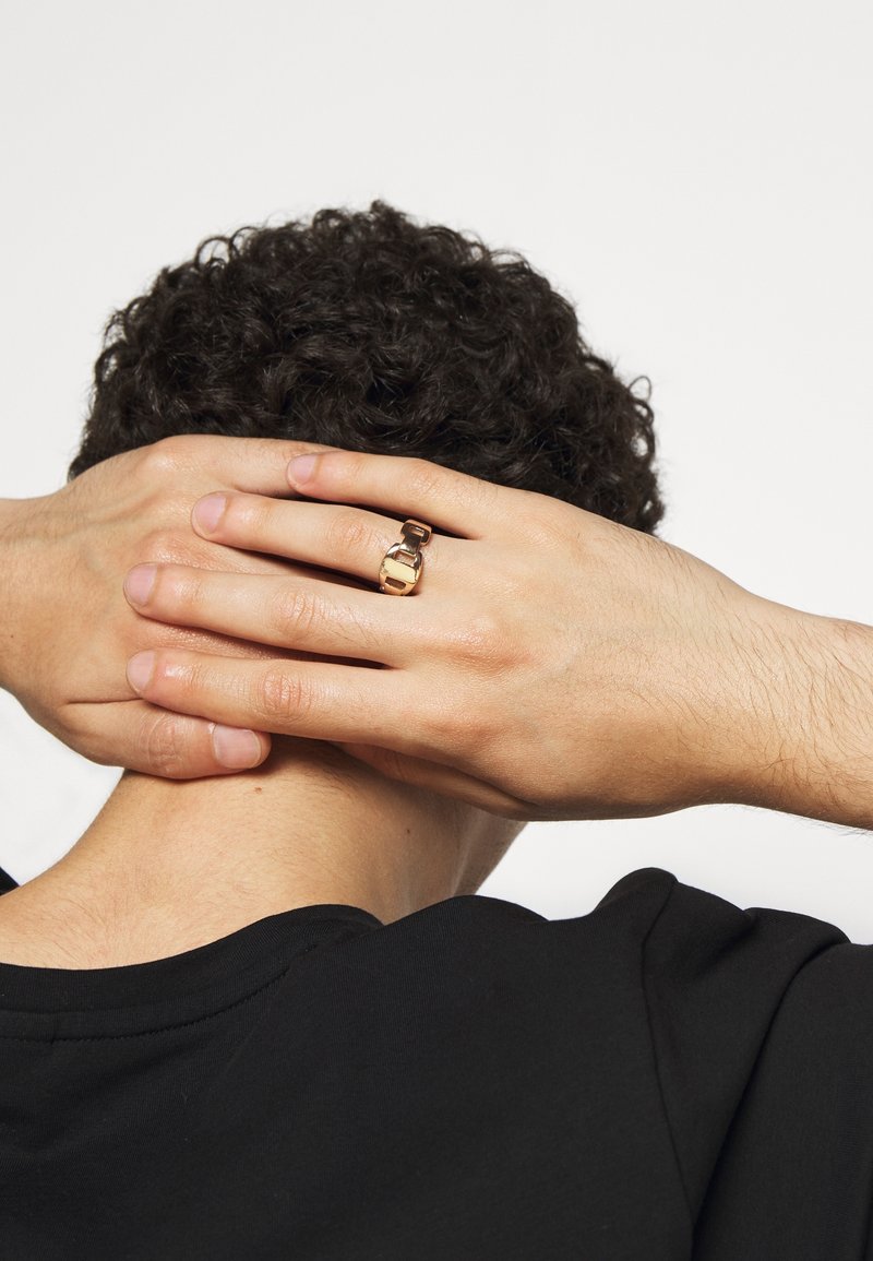 Personne aux cheveux bouclés et foncés tenant l'arrière de la tête, montrant une bague en or avec un motif de boucle au majeur gauche, portant une chemise noire.