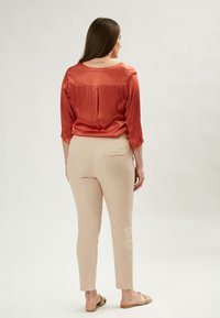 Blusa di satin rame con maniche a tre quarti, dettaglio della piega sul retro, abbinata a pantaloni attillati beige chiaro e sandali bassi.