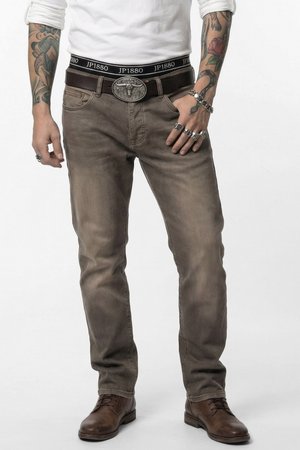 Mann trägt ausgewaschene braune Jeans, braune Lederstiefel, weißes Langarmhemd, schwarzen Gürtel mit silberner Schnalle und mehrere Ringe und Armbänder.