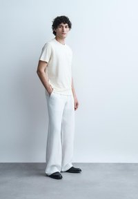 Fiatalember göndör hajjal világos háttér előtt áll, krémszínű rövid ujjú ingben, bő fehér nadrágban és fekete cipőben, kezét a zsebében tartva.