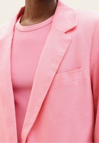 Růžový lněný blazer s klopovým límcem a jednou přední kapsou, nošený přes žebrované růžové tričko, ukazující hladkou texturu a rovnoměrnou barvu.