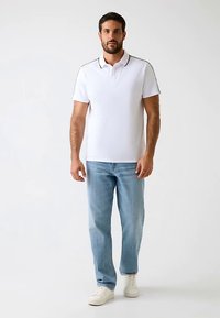 Polo blanc avec des accents noirs, manches courtes, associé à un jean bleu clair et des baskets blanches. Le mannequin est debout, droit, devant un fond neutre.