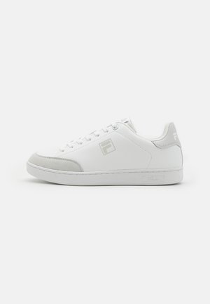 Zapatillas blancas con acentos de ante gris, punta redonda, suela plana y diseño con cordones. Presenta un logo en el lateral y un estilo minimalista.