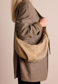 Beige suède schoudertas met een slouchy ontwerp, voorzien van een ritssluiting en leren schouderband. Gepaard met een gestructureerde bruine blazer.