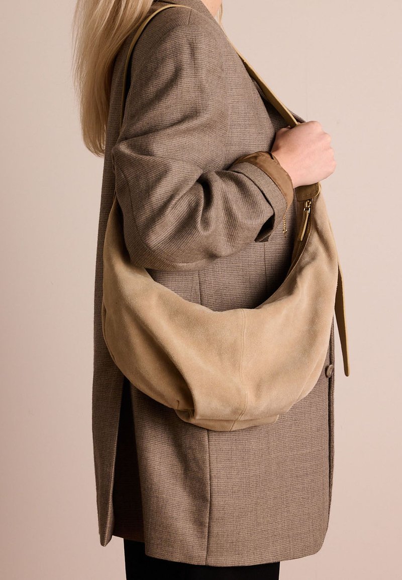 Beige suède schoudertas met een slouchy ontwerp, voorzien van een ritssluiting en leren schouderband. Gepaard met een gestructureerde bruine blazer.