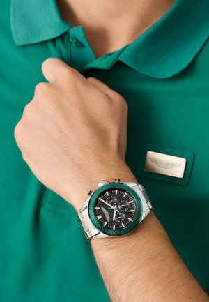 Orologio cronografo argento con lunetta turchese e quadrante nero. Indossato al polso accanto a una maglietta polo verde con una toppa logo.