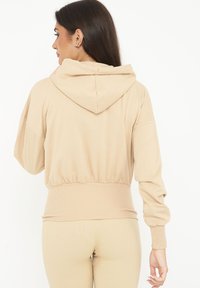 Sudadera corta beige hecha de tela suave, con capucha, bajo acanalado y puños ajustados. Textura suave con una silueta relajada.