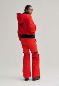 Piros rövidített kapucnis dzseki és hozzáillő, fekete részletekkel díszített, bővülő szárú nadrágok, texturált felülettel és testhezálló derékpánttal.