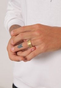 Gold-colored metal ring with a hammered texture, worn on a finger. Hands have visible tattoos and slight discoloration. Background is neutral.