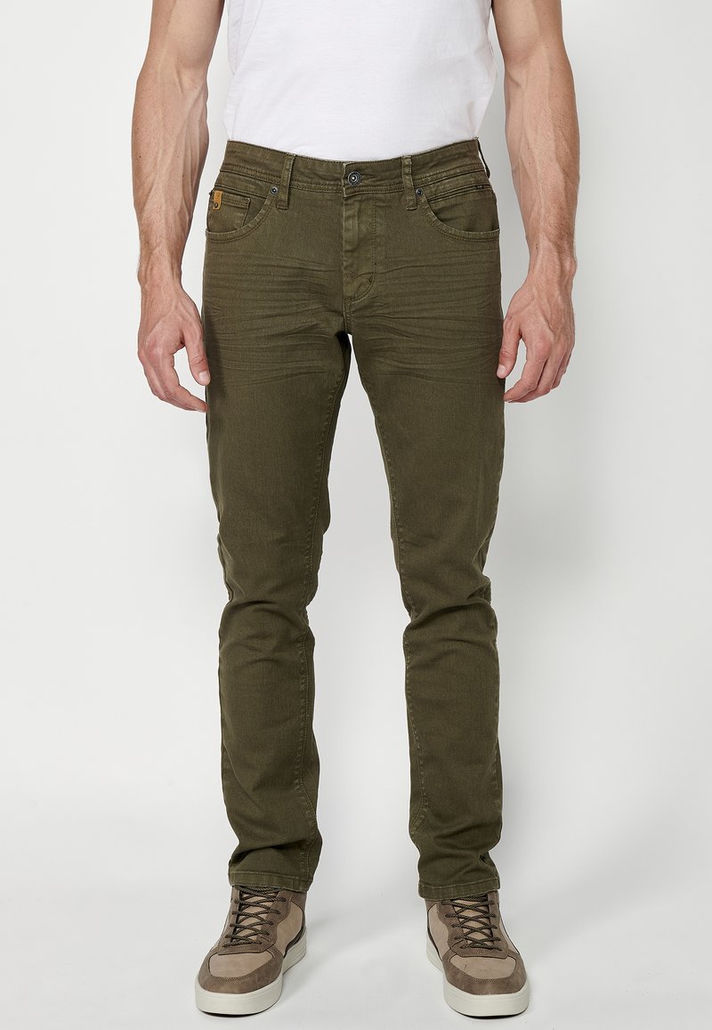 Pantalones ajustados de color verde oliva con una textura suave, cinco bolsillos, cierre de botón y costuras tonales; combinados con zapatillas beige.