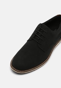 Chaussure de ville noire à lacets avec dessus lisse, semelle cousue marron et blanche, vue en angle supérieur latéral sur fond gris.