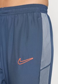 Marine atletiske shorts med elastisk talje, mesh-sider og et orange Nike-logo på venstre forkant. Glat stofstruktur.