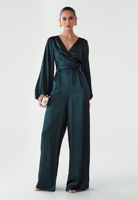 Grüner Satinoverall mit tiefem V-Ausschnitt, langen Ballonärmeln und weitem Bein. Verfügt über eine taillierte Passform und ein dezentes strukturiertes Muster.
