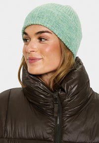 Gorro de punto acanalado verde claro, llevado con una chaqueta de plumas marrón con cuello alto y cierre de cremallera. Textura suave evidente.