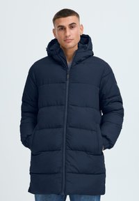 Marineblå pufferjakke med hette, med quiltet design, frontlynlås og side-lommer. Materialet ser mykt og isolert ut.