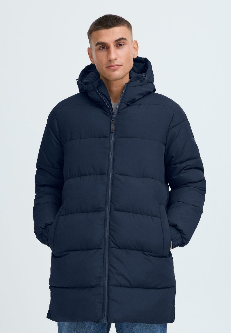 Marineblå pufferjakke med hette, med quiltet design, frontlynlås og side-lommer. Materialet ser mykt og isolert ut.