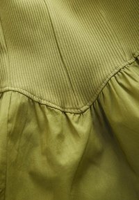 Le tissu en maille côtelée passe à un tissu froncé lisse en vert olive, montrant des coutures détaillées et un contraste de texture.