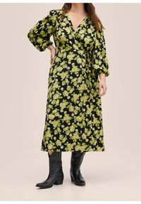 Robe portefeuille noire à fleurs avec des fleurs vertes et crème, manches bouffantes longues et une taille nouée. Associée à des bottes de cowboy noires.