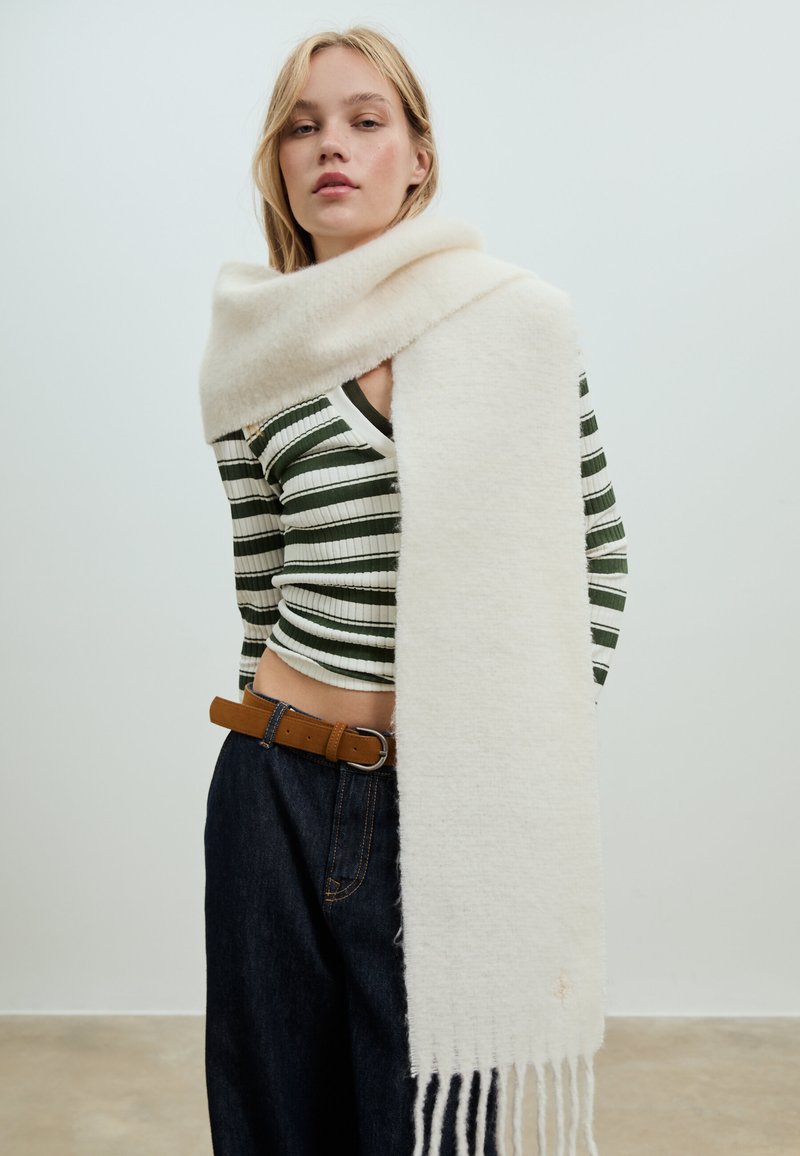 Zachte witte sjaal van wol, met een gladde textuur, franjes aan de randen, nonchalant gedrapeerd over een persoon in een gestreept shirt en spijkerbroek.