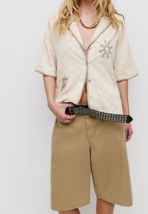 Chemise crème à manches courtes avec broderie florale, associée à un short kaki. Ceinture noire à clous soulignant la taille.