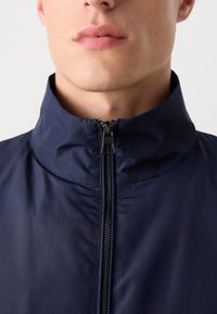 Navy blue lightweight jacket featuring a high collar and a central zipper. The fabric has a smooth, matte texture with minimal stitching details.