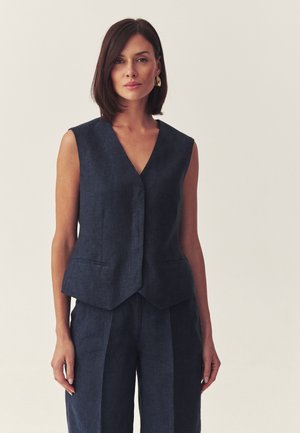 Kobieta o ramionach długości brązowych włosach, ubrana w bezrękawnik w ciemnoniebieskim kolorze i pasujące spodnie, stojąca na tle jasnego, jednolitego tła.