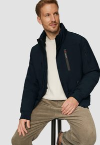 Marineblauwe gewatteerde jas met hoge kraag, een ritssluiting aan de voorkant en zijzakken. Draagt over een crèmekleurige trui en gecombineerd met beige corduroy broek.