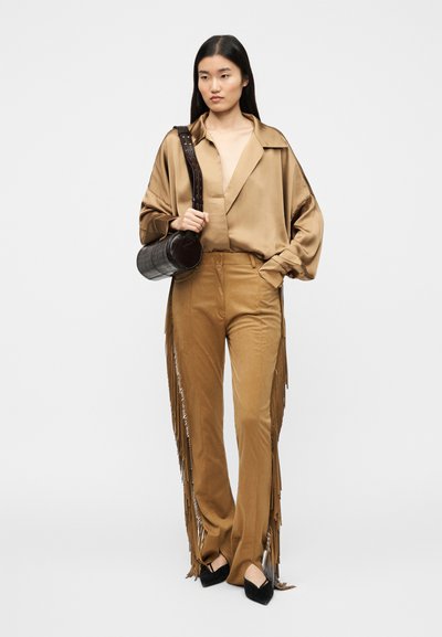 Μπλούζα από μπρονζέ σατέν με βαθύ V-λαιμόκοψη, συνδυασμένη με καφέ παντελόνια με φούντες. Το μοντέλο κρατάει μια στρογγυλή τσάντα με σκούρο κροκοδειλένιο μοτίβο. Μαύρα παπούτσια με τακούνι.