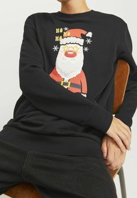 Una persona che indossa una felpa nera con un grafico colorato di Babbo Natale e il testo "Ho Ho Ho", seduta su una sedia di legno.