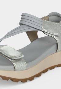 Sandalia plateada con punta abierta, con tiras ajustables de velcro, plantilla acolchada y suela beige con suela exterior marrón texturizada.