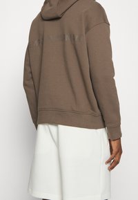 Brown hooded sweatshirt with a relaxed fit, featuring a textured logo print on the back, paired with light-colored shorts.