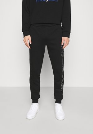 Mann trägt schwarze Emporio Armani Jogginghose mit Logotext am linken Bein und weiße Sneakers vor schlichtem Hintergrund.